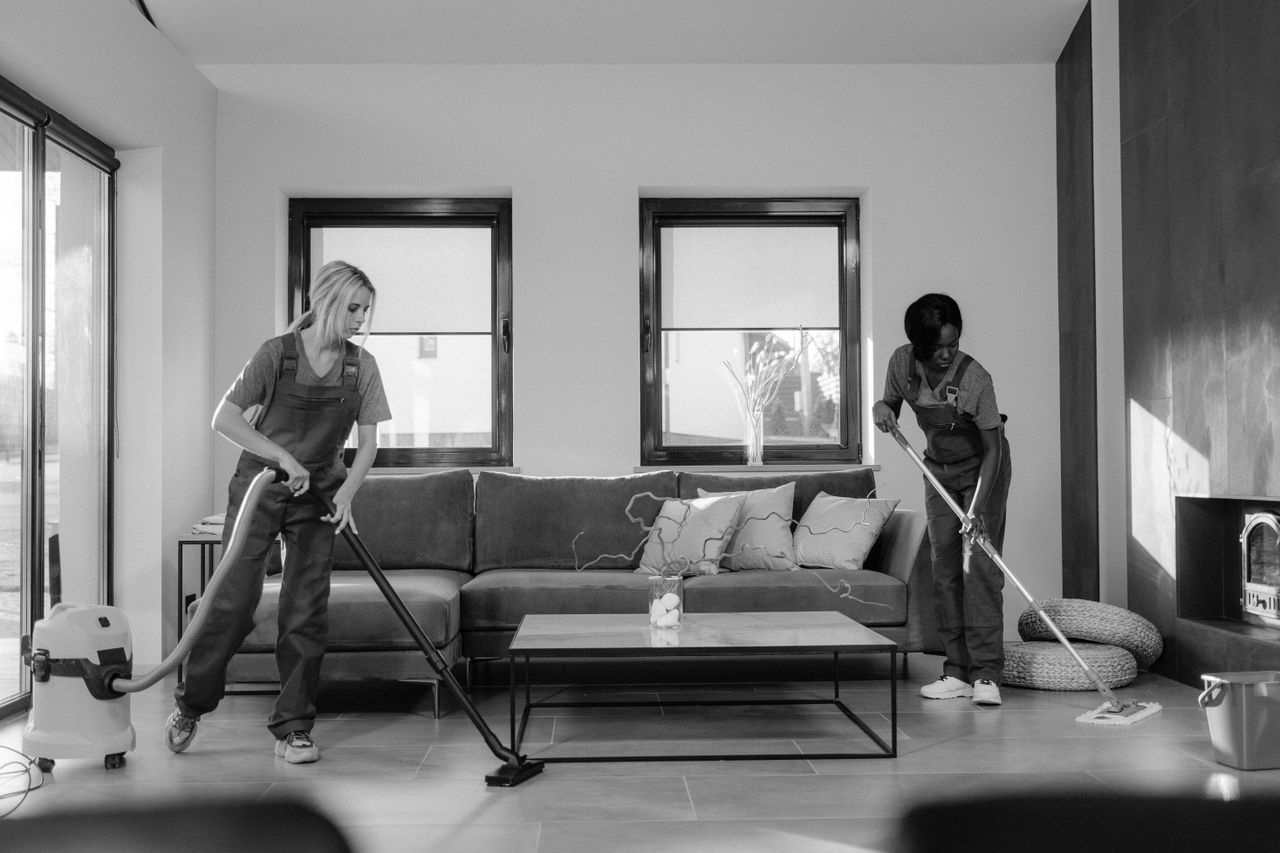 hero-img-02 Two women in work clothes cleaning a modern living room using a vacuum and mop.